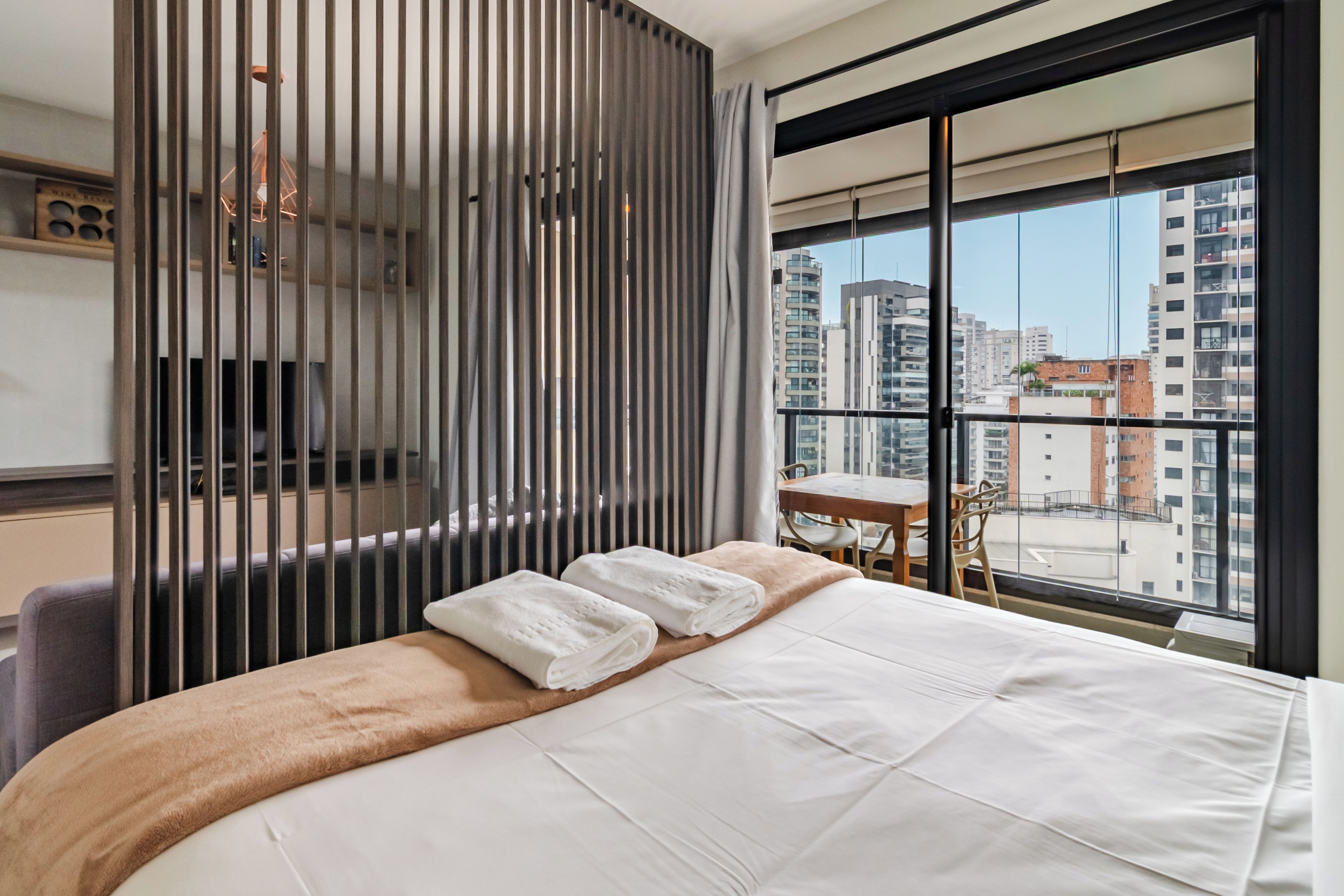 A hotel room with a large bed, a desk, and a view of the cityscape through floor-to-ceiling windows.