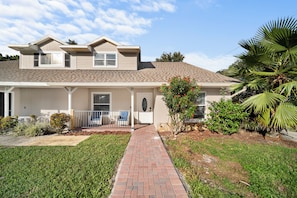 A two-story house with a front porch, a brick pathway leading to the door, and a small garden area with various plants and a palm tree. - Heated Saltwater Pool, Pac-Man Arcade, 2 Putting Greens & Tampa Beach Getaway (Tampa)