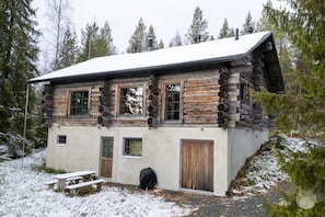 Eine Blockhütte mit schneebedecktem Dach, einer Holztür und einem Picknicktisch in einem verschneiten Wald.