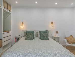 A bedroom with a large bed, two nightstands, a white chair, and a shelving unit. - CHL La Palma Beach, infront of the sea (Santa Cruz de La Palma)
