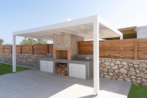 An outdoor kitchen area with a stone wall, wooden fence, and a white pergola structure. - Serenity Villa Thalassa (Kolympia)