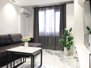 Ein modernes Wohnzimmer mit einem dunklen Sofa, einem weißen Couchtisch, einer Topfpflanze und einem großen Fenster mit Vorhängen.