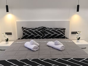Ein modernes Schlafzimmer mit einer grauen gesteppten Bettdecke, zwei schwarz-weißen Kissen mit Muster, zwei aufgerollten Handtüchern und zwei weißen Blumenvasen auf Nachttischen.