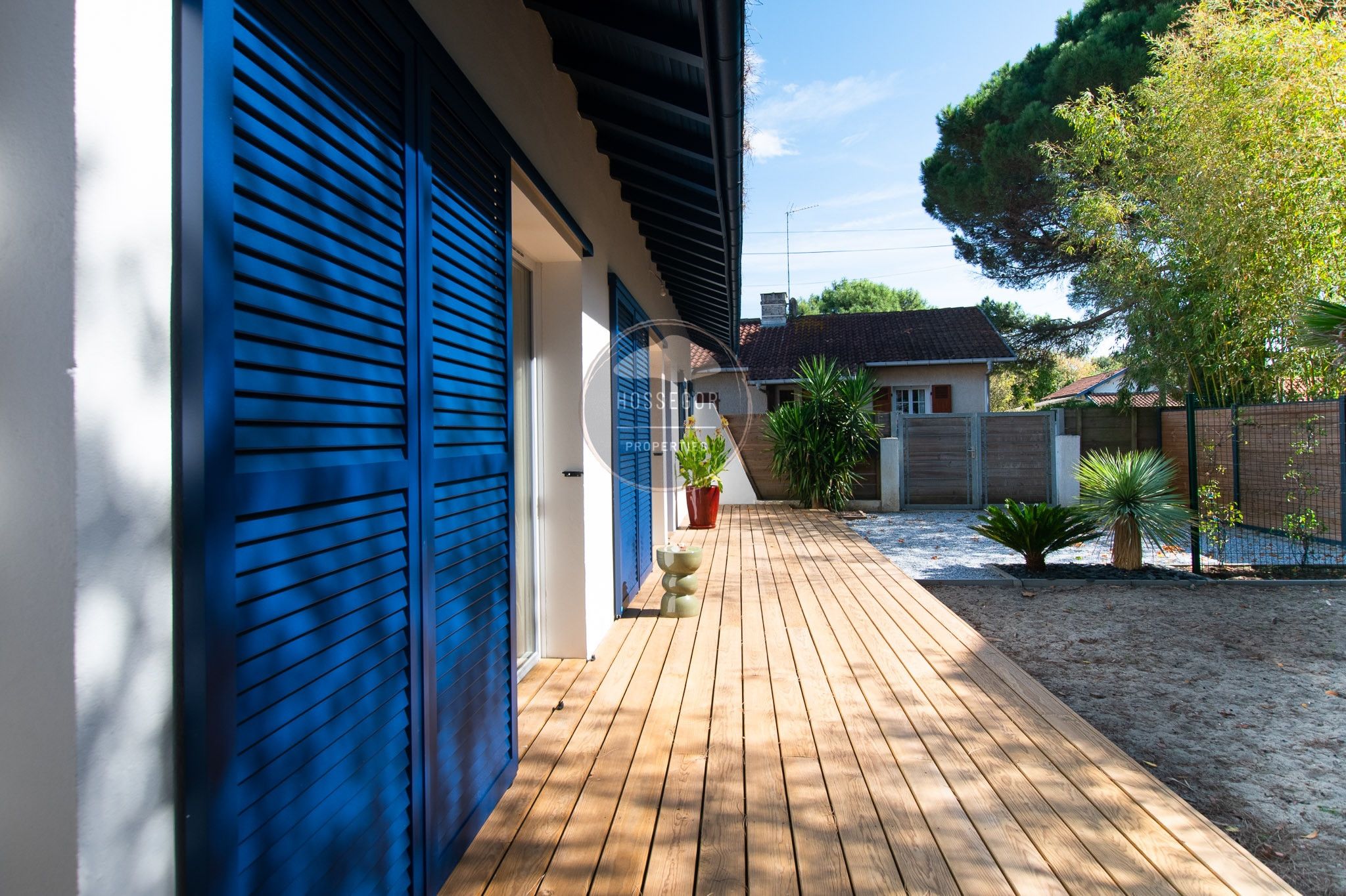 A wooden deck leading to a building with blue shutters, a small garden with plants, and a fence.