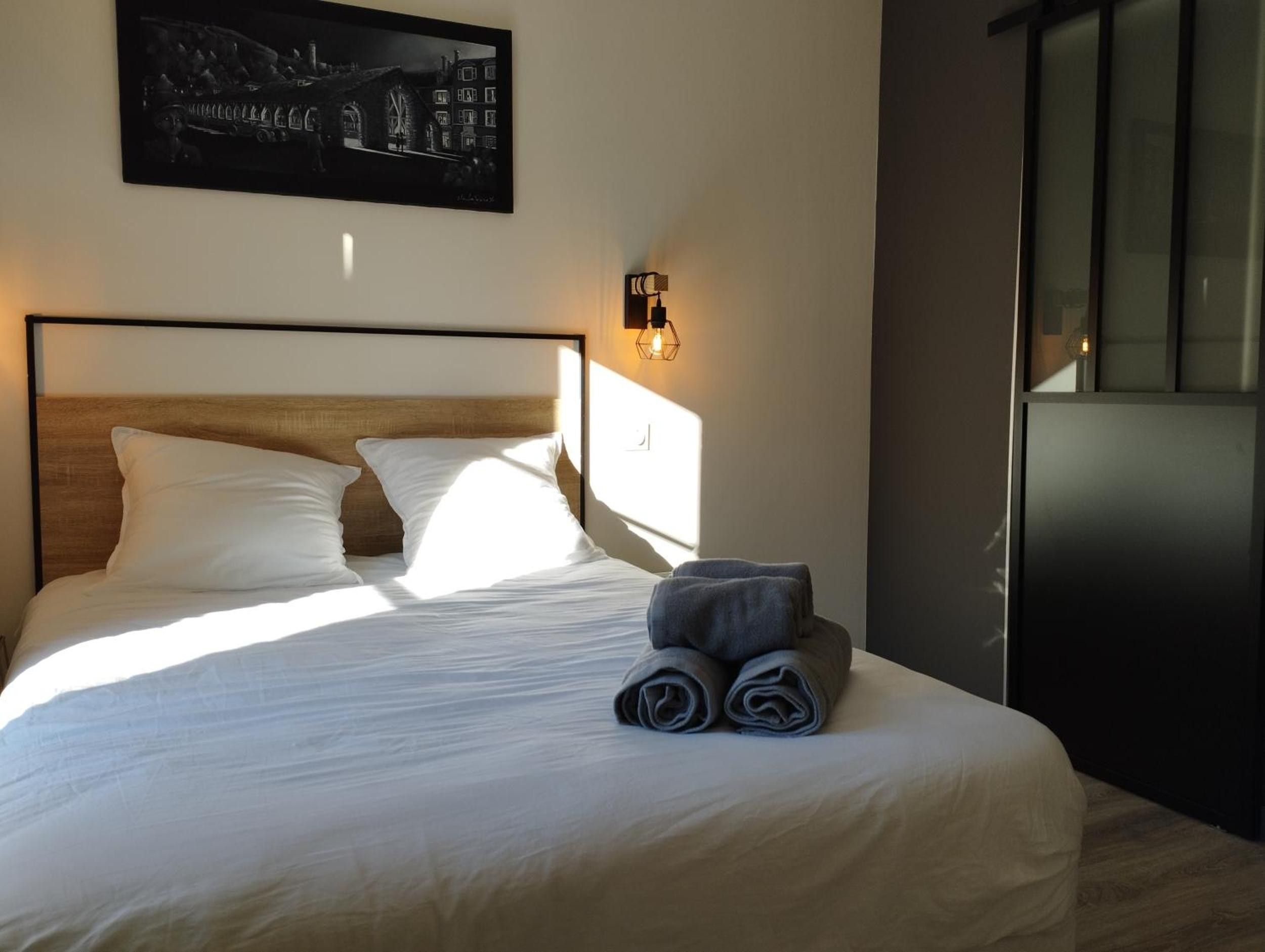 A neatly made bed with white linens and two pillows, a folded towel on the side, a wall-mounted light fixture, and a framed picture above the bed.