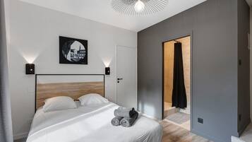 A modern bedroom with a wooden headboard, a bed with white linens, a wall-mounted light fixture, and a framed picture.