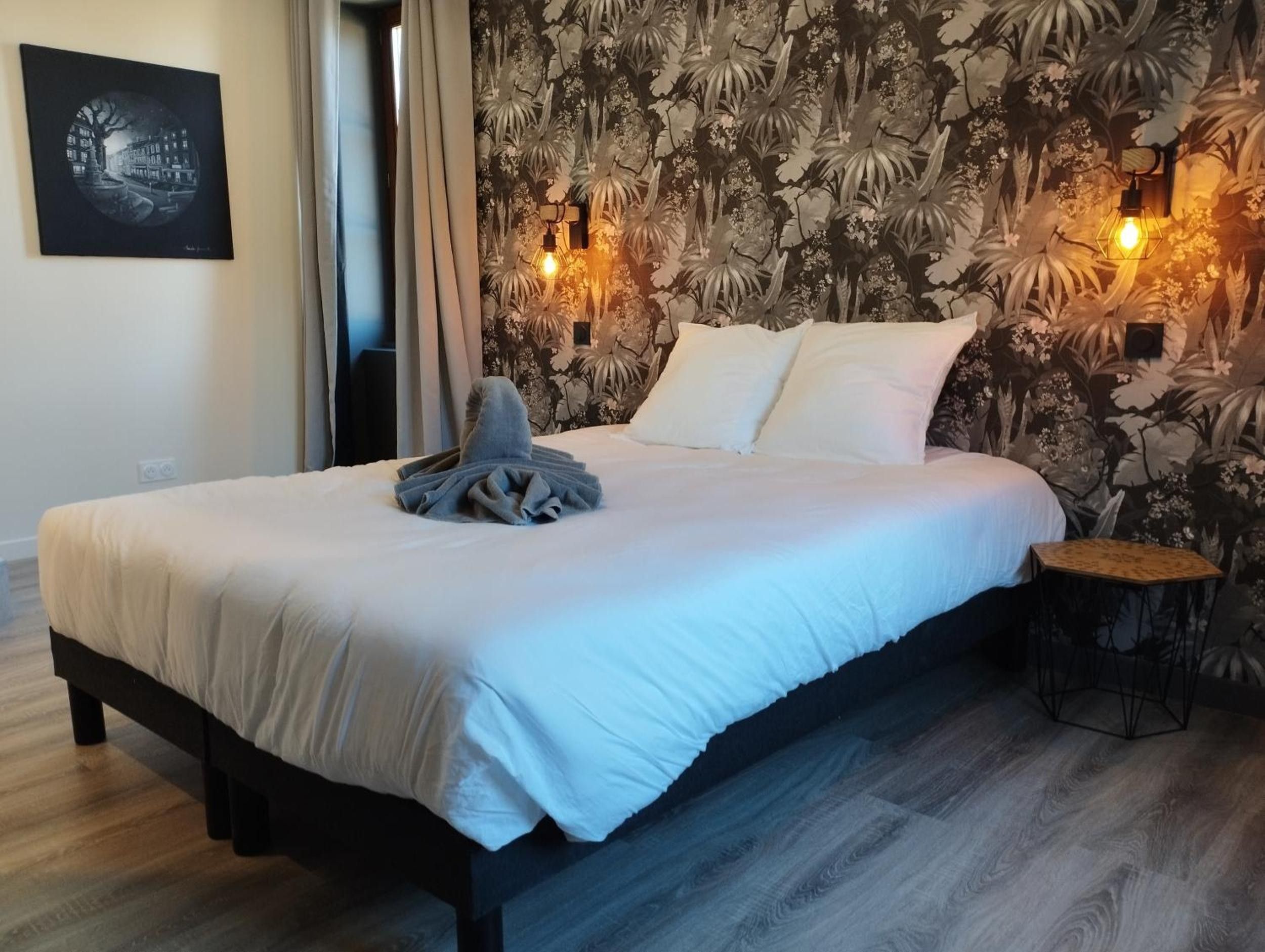 Una cama bien tendida con edredón blanco, una toalla doblada y dos almohadas. Hay una lámpara de pared en la pared detrás de la cama.
