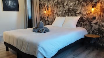 Una cama bien tendida con edredón blanco, una toalla doblada y dos almohadas. Hay una lámpara de pared en la pared detrás de la cama.