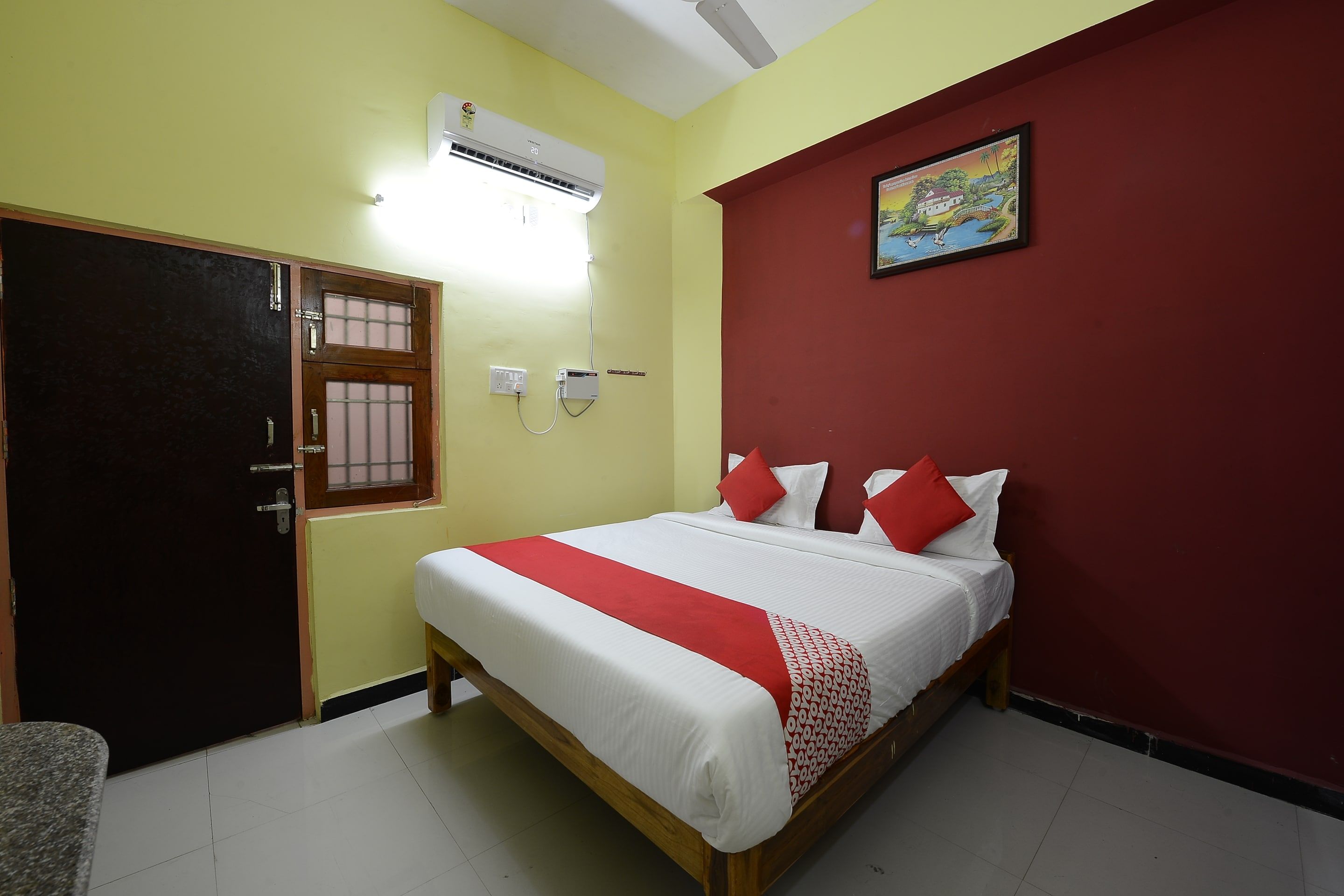 A hotel room with a bed, a door, an air conditioner, and a wall-mounted electrical outlet.