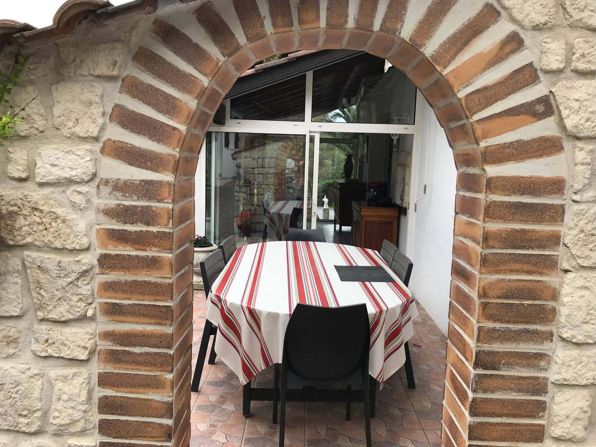 Un espace extérieur aménagé pour les repas, avec une arche en briques, une table recouverte d’une nappe à rayures et des chaises.