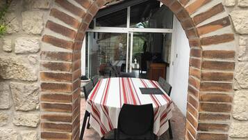 Un espace extérieur aménagé pour les repas, avec une arche en briques, une table recouverte d’une nappe à rayures et des chaises.