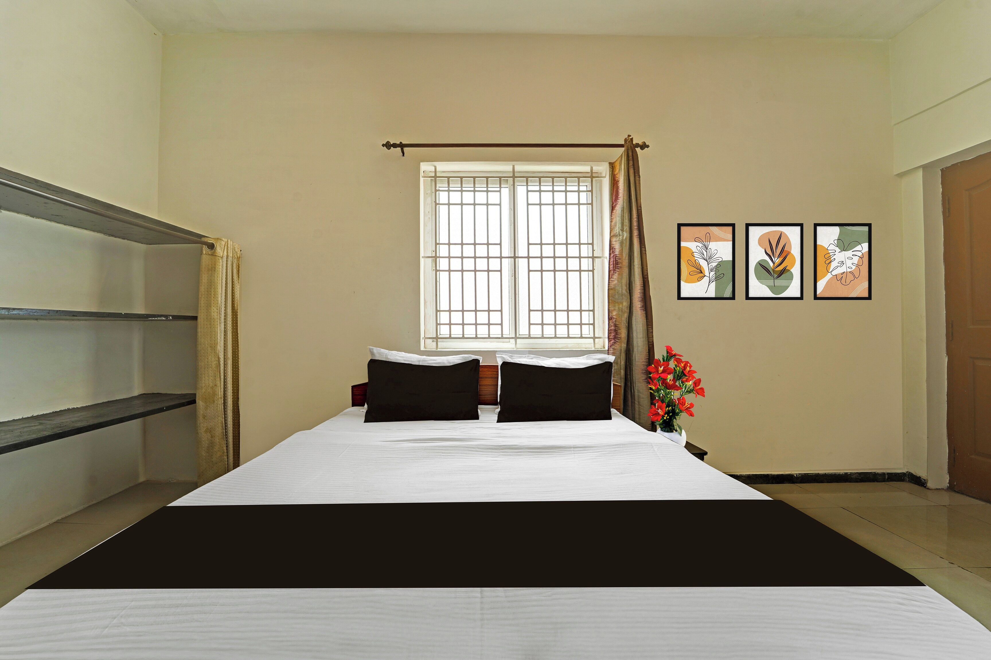 Une chambre d’hôtel avec un lit, une fenêtre avec un rideau, une étagère fixée au mur et trois tableaux encadrés.