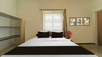 Une chambre d’hôtel avec un lit, une fenêtre avec un rideau, une étagère fixée au mur et trois tableaux encadrés.