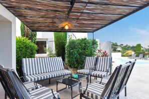 A covered outdoor seating area with striped lounge chairs and a table, surrounded by greenery and a view of a swimming pool. - Villa La Chantyviere - Piscine Privée, Pool House (Sainte-Maxime)