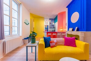 Ein helles Interieur mit einem gelben Sofa, einem kleinen Tisch mit einer Pflanze und einer Küchenzeile im Hintergrund.