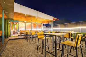 An outdoor seating area with tables and chairs, a wooden ceiling, and a building with a glass facade in the background.