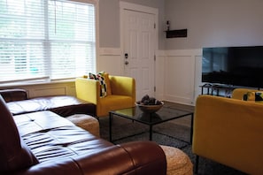 Een woonkamer met een gele bank, een bruine leren zithoek, een glazen salontafel en een flatscreen televisie.