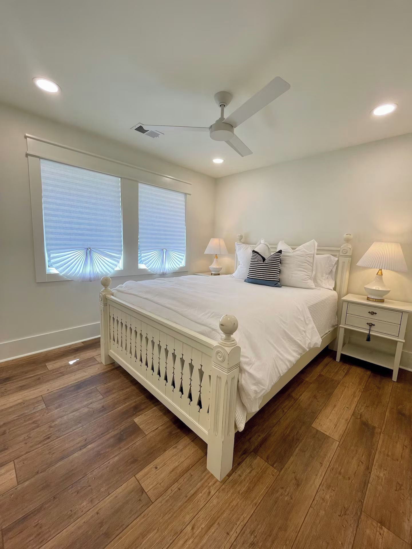 A bedroom with a white bed, two bedside tables, a ceiling fan, and a window with blinds.