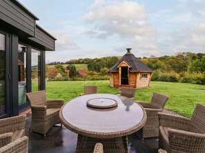 A patio with a round table and wicker chairs, overlooking a grassy area and a small wooden structure. - Hanbury House (Ross-on-Wye)