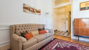 Ein Wohnzimmer mit beiger Couch, gemusterten Kissen, einem rot-weißen Teppich, einem Bild an der Wand und einem Holzschrank.