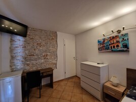 Ein kleiner, kompakter Raum mit einer Steinswand, einem weißen Schrank, einem Schreibtisch, einem Stuhl, einem Bett mit einem sternenmuster-Kissen und einer Wandlampe.