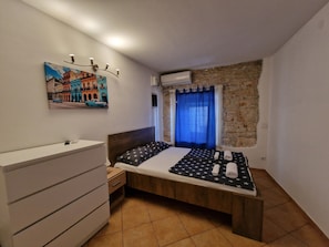 A bedroom with a bed, a dresser, a painting, a window with curtains, and an air conditioning unit. - Damian (Pula)