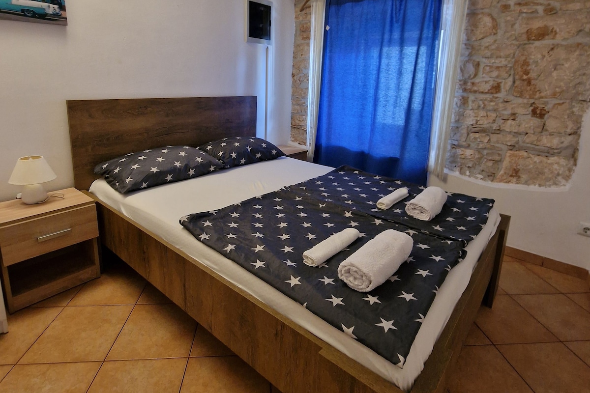 A bedroom with a bed, a nightstand, a lamp, a wall-mounted air conditioner, and a window with a curtain.