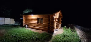 Une cabane en bois avec une seule fenêtre et une porte, entourée d’un mur de pierres et d’une clôture en métal.