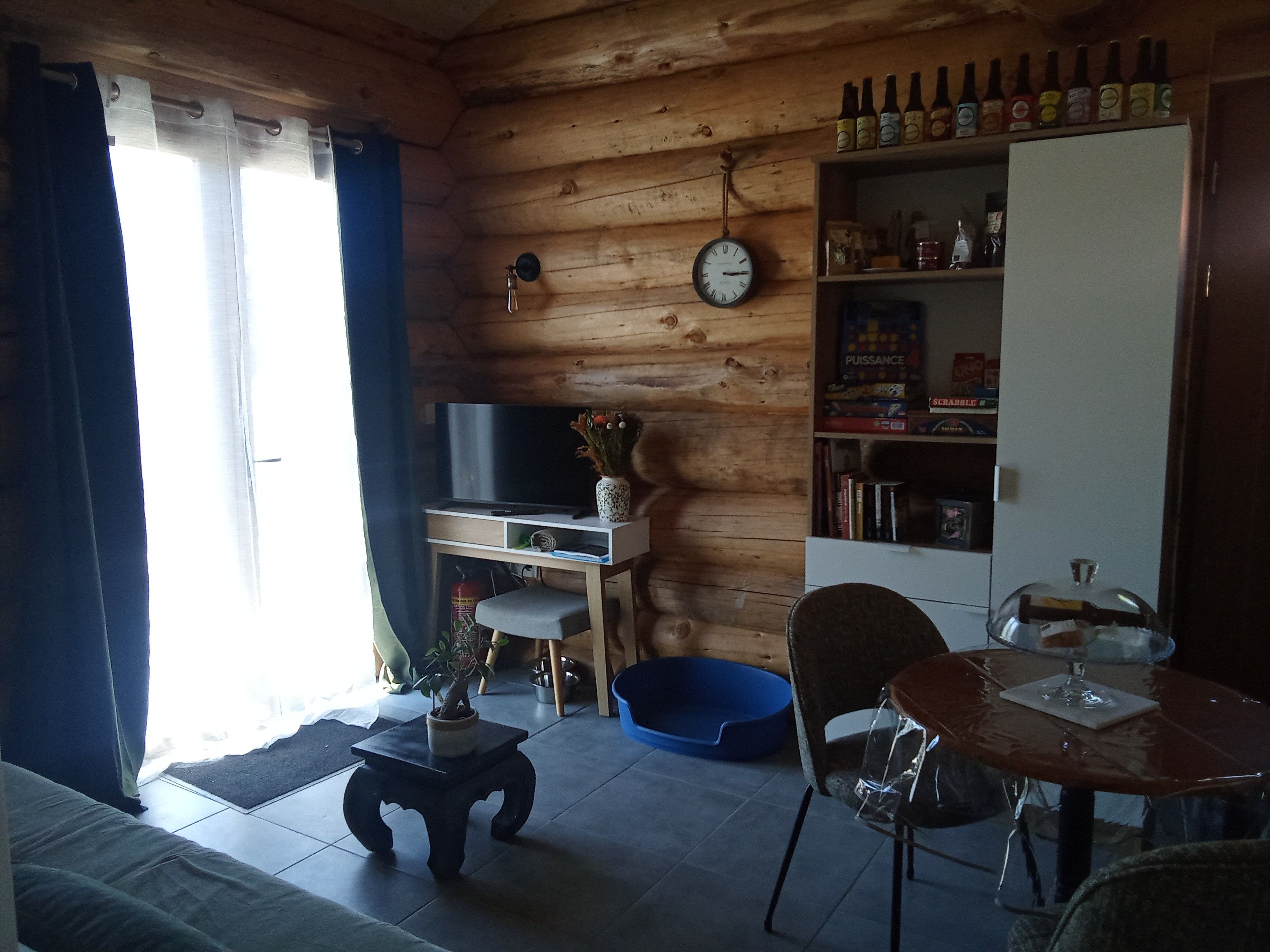 Een gezellige woonkamer met houten inrichting, een flatscreen televisie, een kleine tafel met een vaas en een boekenkast met diverse spullen.