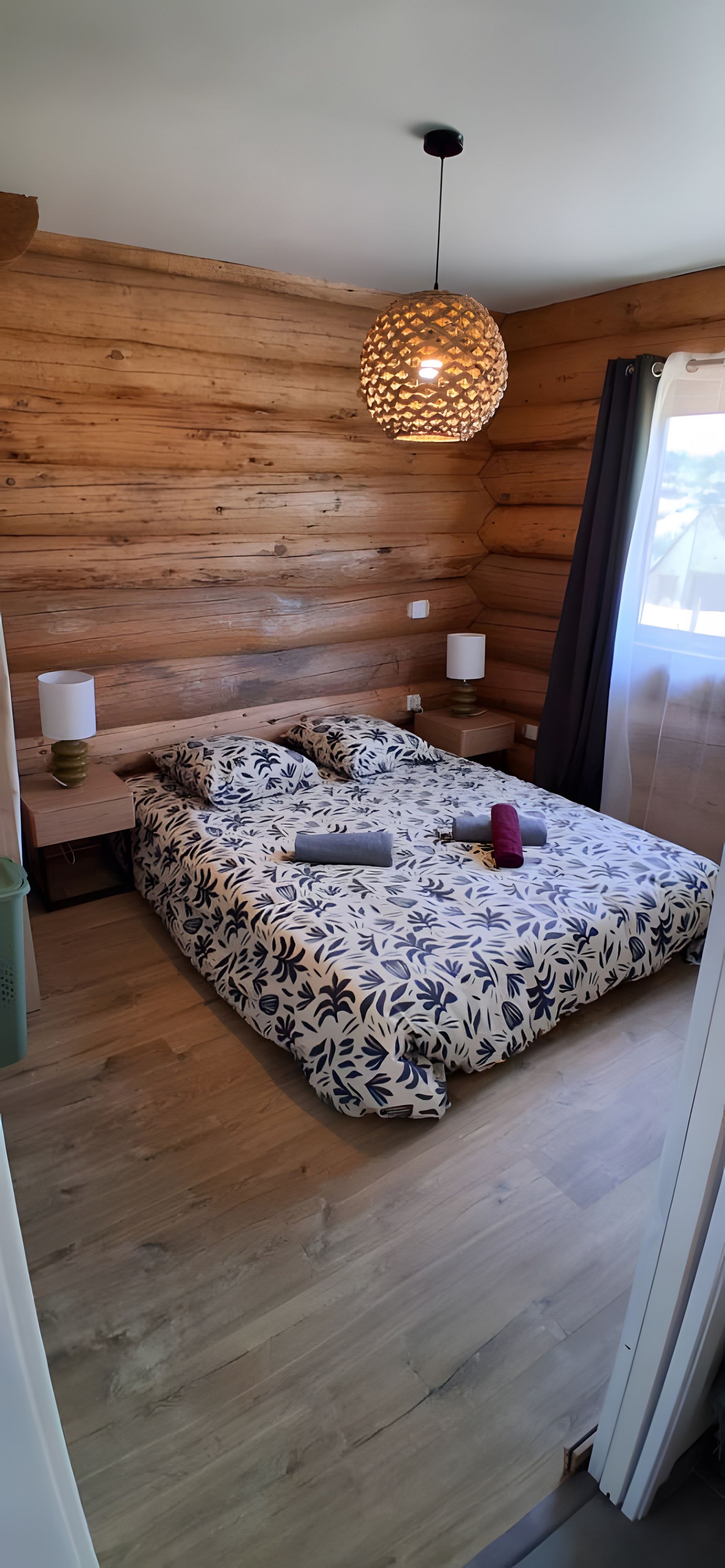 Makuuhuone, jossa on puusäleseinä, kukallinen päiväpeite sängyllä, riippuvalaisin ja ikkuna, jossa on verhot.