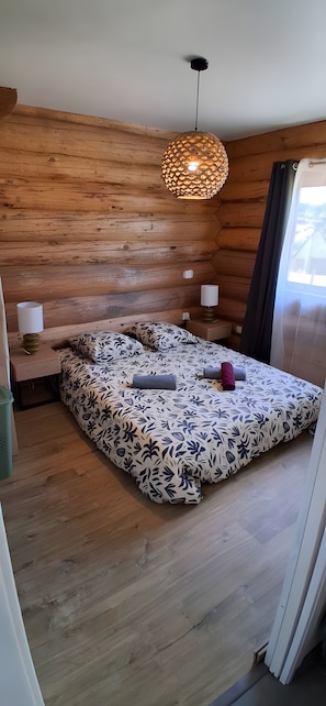 Un dormitorio con pared de listones de madera, cama con colcha estampada, lámpara colgante y ventana con cortinas.
