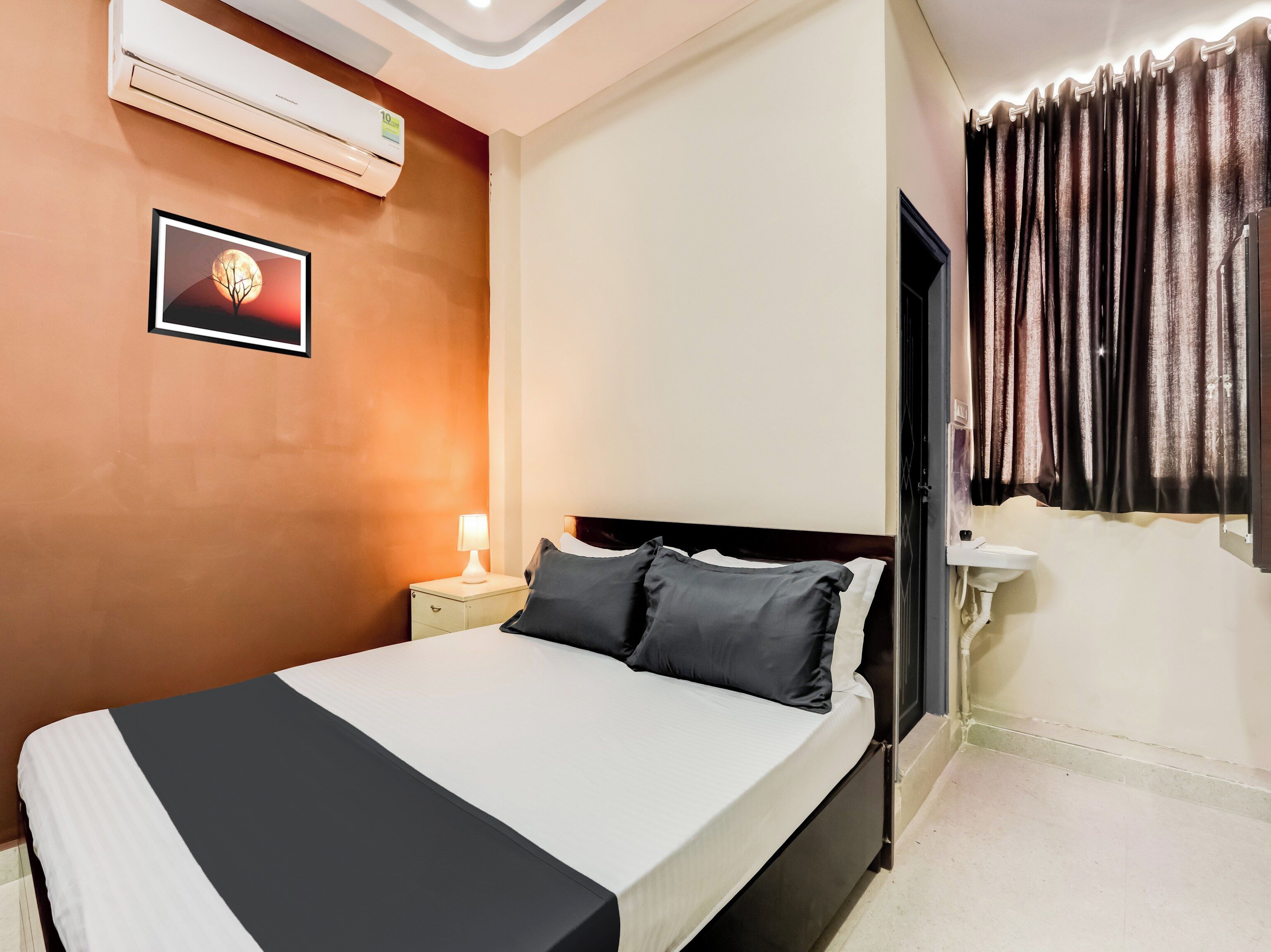 A hotel room with a bed, bedside table, lamp, air conditioning unit, and a bathroom with a sink.