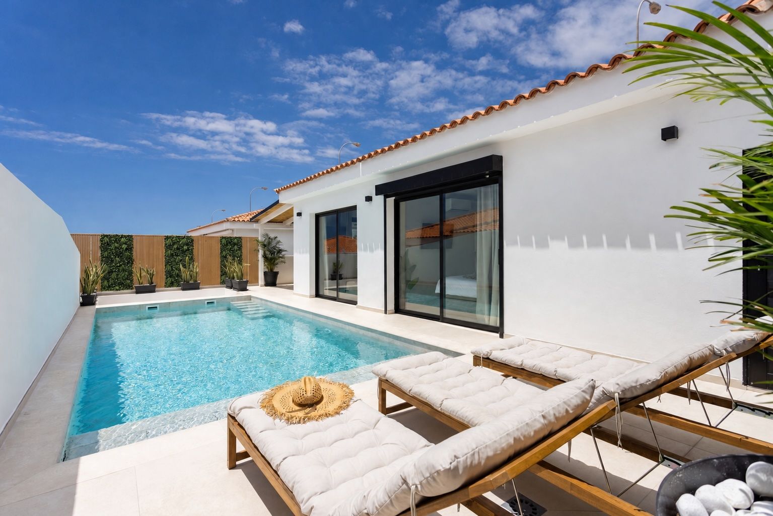 A modern pool area with a clear blue pool, lounge chairs, and a white building with a tiled roof.