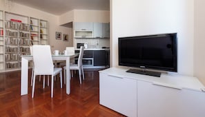 Een moderne woonkamer met een flatscreen televisie, witte meubels, een boekenkast en een keukenhoek.