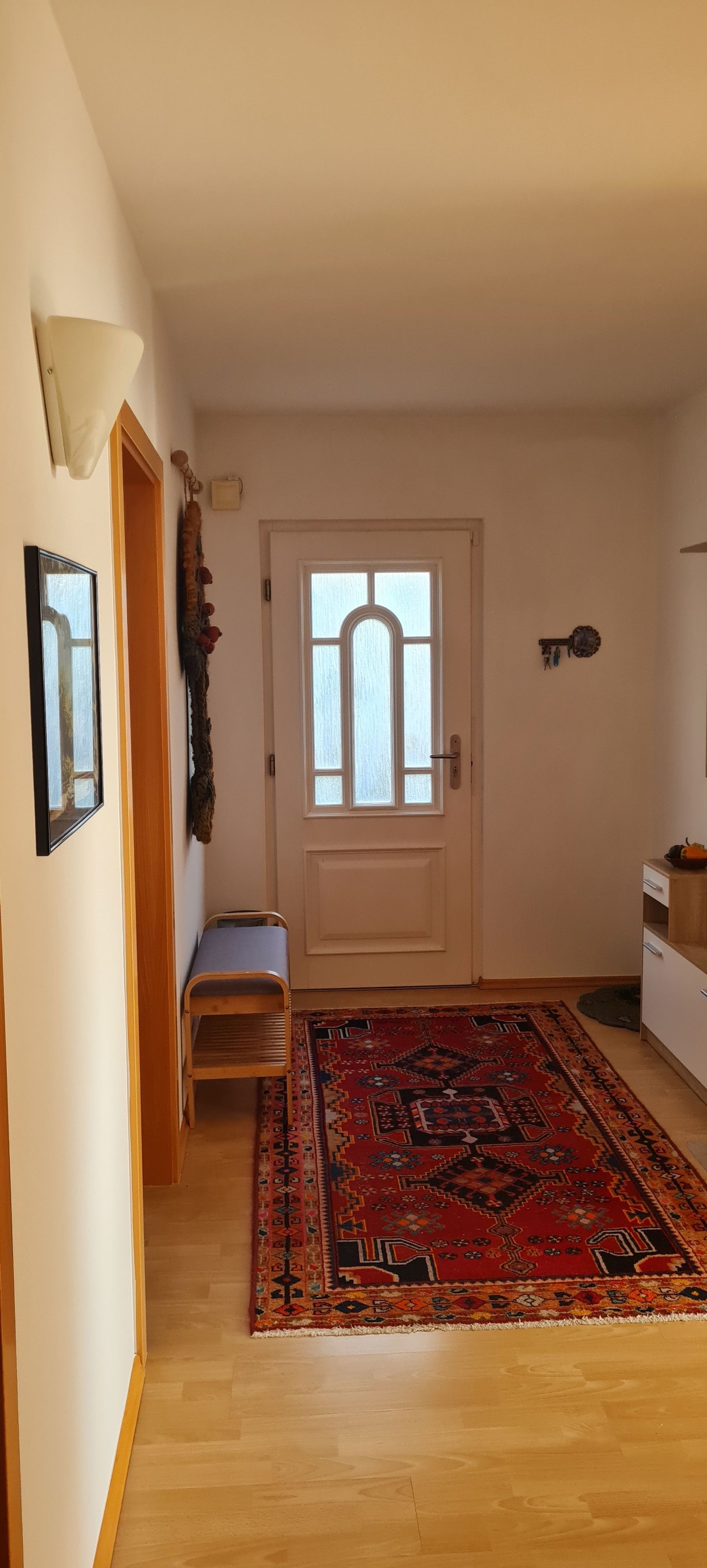 A hallway with a wooden bench, a patterned rug, a white door with a window, and a small shelf with decorative items.