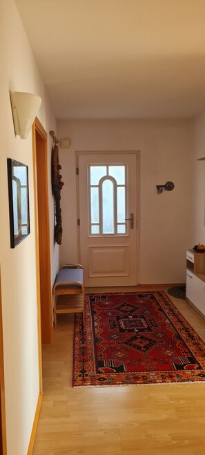 Un couloir avec un banc en bois, un tapis à motifs, une porte blanche avec une fenêtre et une petite étagère ornée d’objets décoratifs.