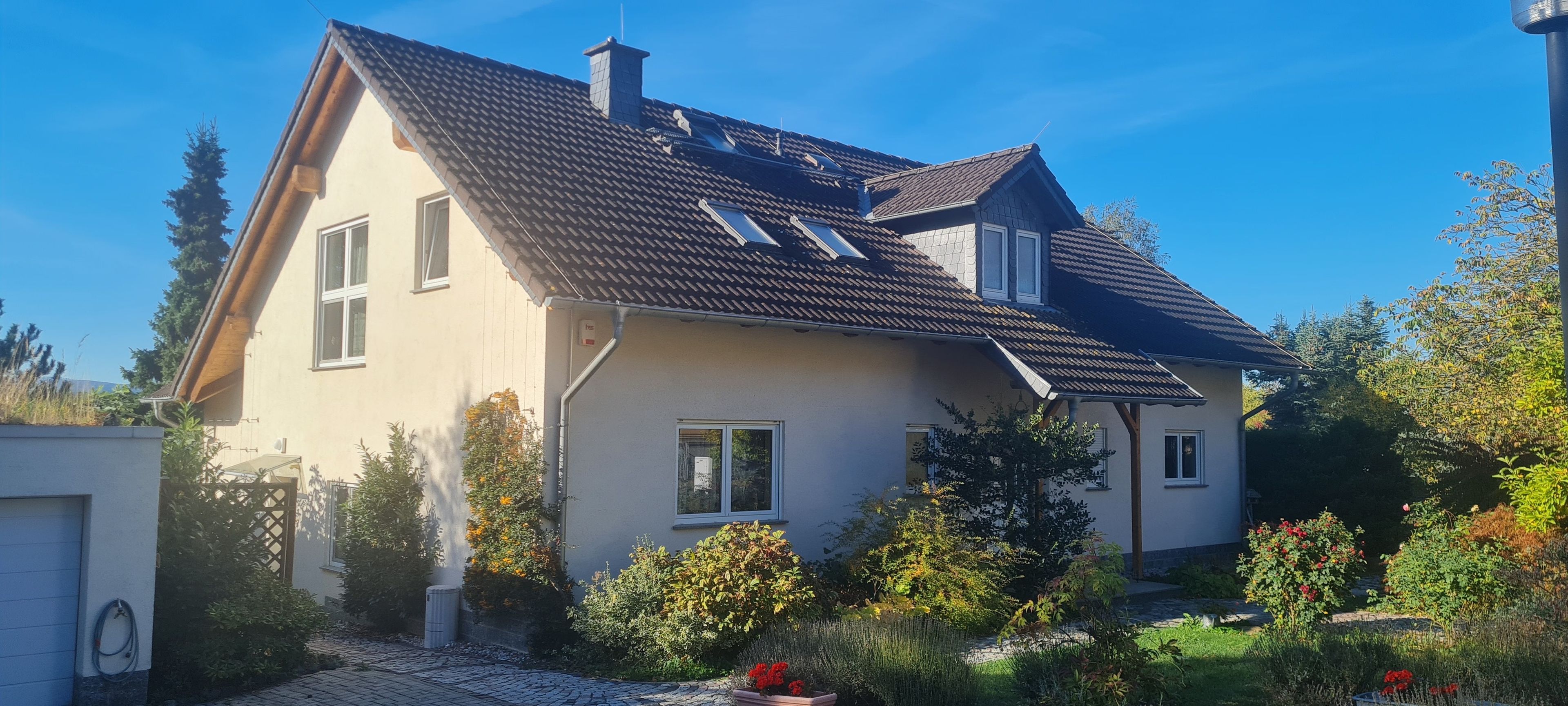 Ein zweigeschossiges Haus mit geneigtem Dach, zahlreichen Fenstern und einem Garten mit verschiedenen Pflanzen und Blumen.