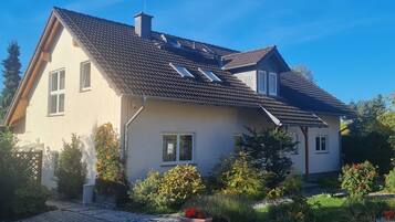 Ein zweigeschossiges Haus mit geneigtem Dach, zahlreichen Fenstern und einem Garten mit verschiedenen Pflanzen und Blumen.
