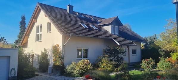 Ein zweigeschossiges Haus mit geneigtem Dach, zahlreichen Fenstern und einem Garten mit verschiedenen Pflanzen und Blumen.