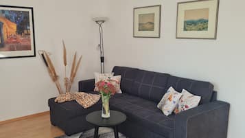 A living room with a dark blue sofa, a small round table, and a tall floor lamp. There are three framed artworks on the wall.
