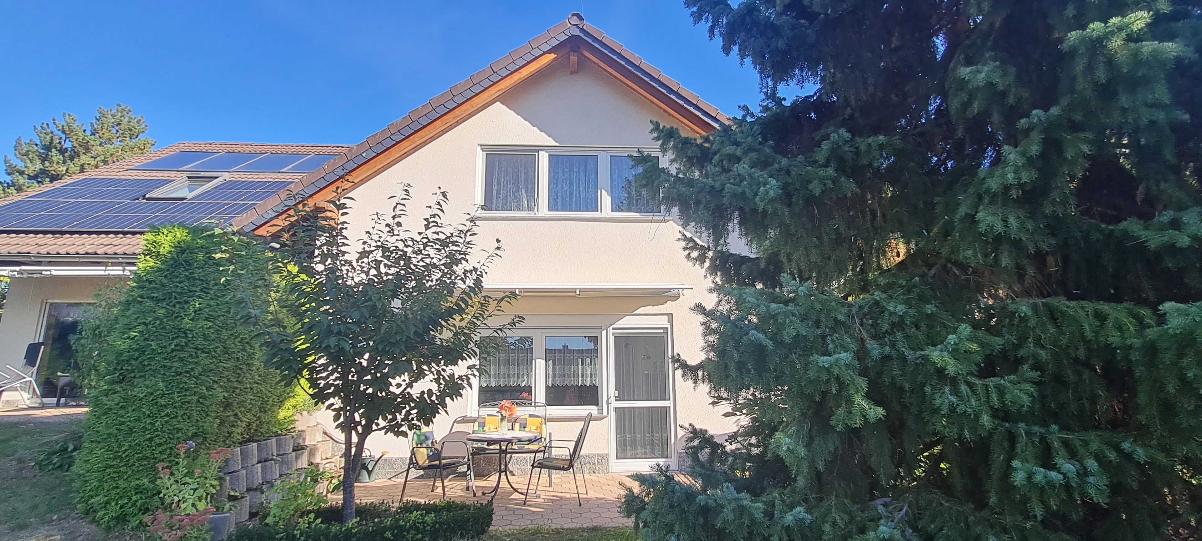 Ein zweistöckiges Haus mit einer Solaranlage auf dem Dach, einer Veranda mit Tisch und Stühlen und einem großen immergrünen Baum zur Rechten.