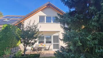 Ein zweistöckiges Haus mit einer Solaranlage auf dem Dach, einer Veranda mit Tisch und Stühlen und einem großen immergrünen Baum zur Rechten.