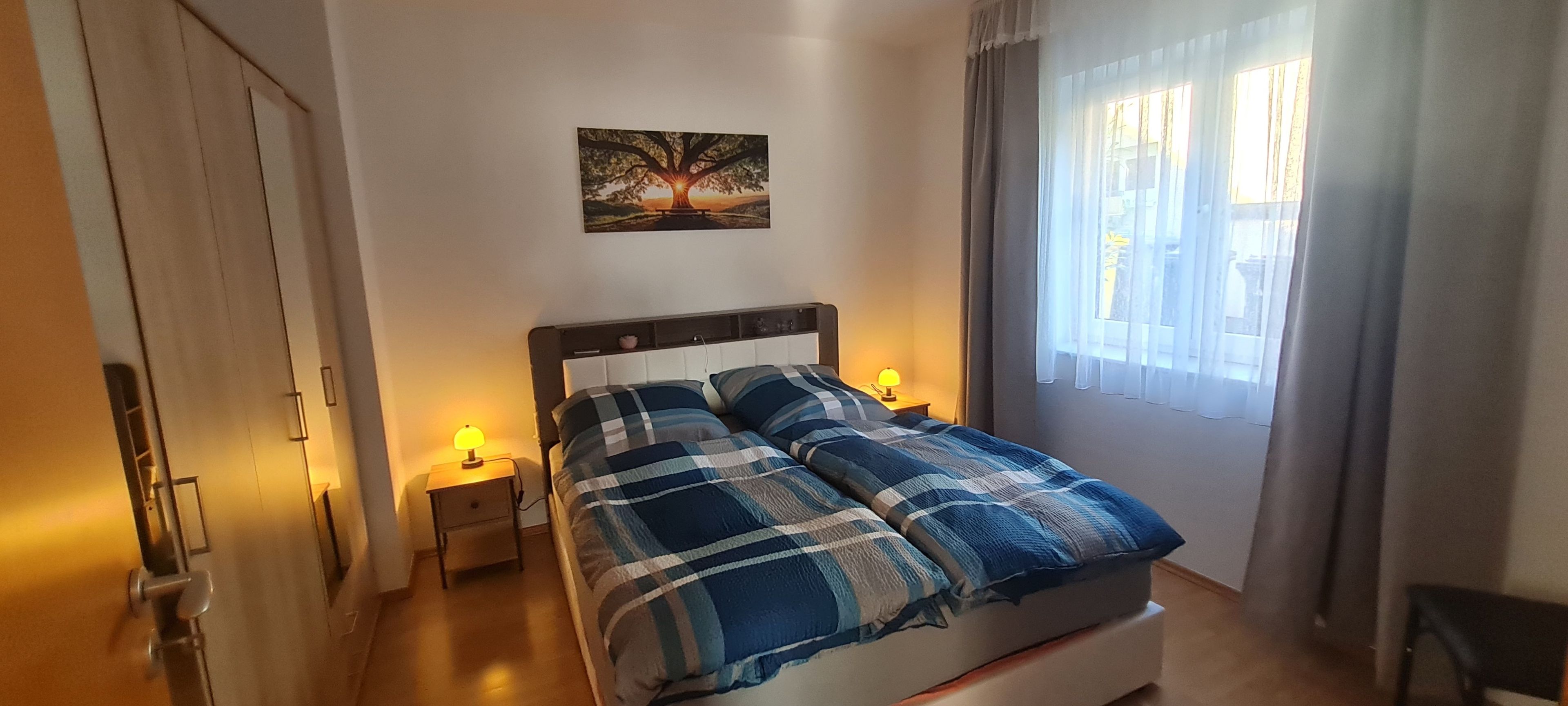 Ein Schlafzimmer mit einem Bett, zwei Nachttischlampen, einem Wandbild und einem Fenster mit Vorhängen.