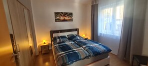 Ein Schlafzimmer mit einem Bett, zwei Nachttischlampen, einem Wandbild und einem Fenster mit Vorhängen.