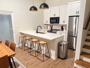 A modern kitchen with white cabinets, a stainless steel refrigerator, a microwave, and a sink. There are four stools around the island and a trash can. - Golf Club Hideaway (Oxford)