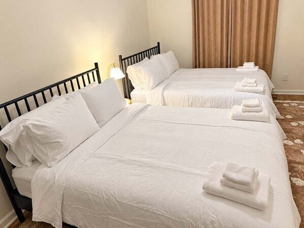 Habitación de hotel con dos camas, cada una con ropa blanca y almohadas, y una mesita de noche con lámpara.