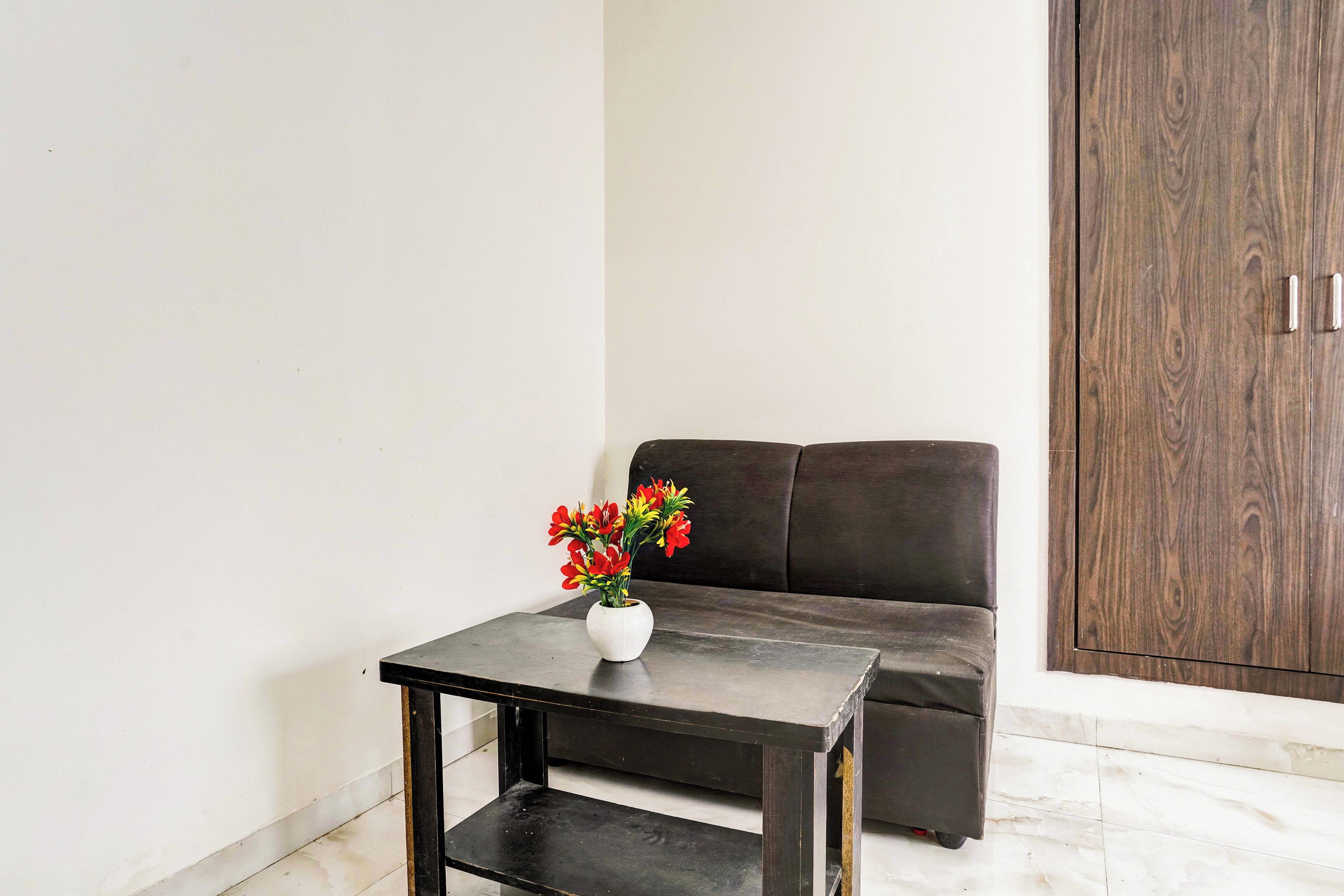 Uma pequena mesa com um vaso branco contendo flores vermelhas, colocada em frente a um sofá marrom-escuro e um armário de madeira.