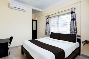 Quarto de hotel com cama, escrivaninha, ar-condicionado, janela com cortinas e porta.