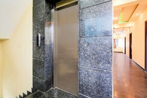Um elevador moderno com acabamento metálico, ao lado de um corredor com piso lustroso e paredes com padrão pontilhado.
