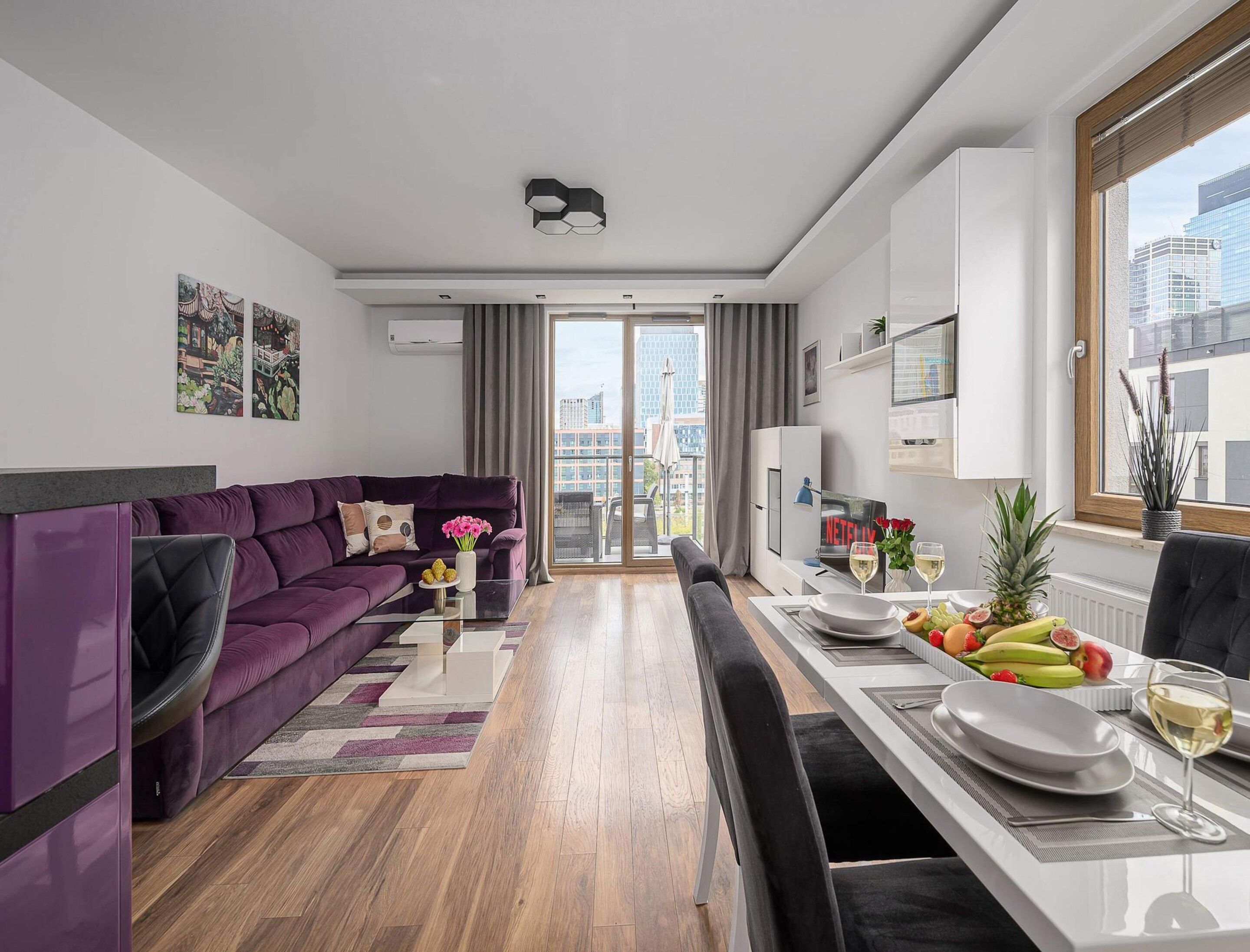 A modern living room with a purple sofa, a dining table set for two, and a city view through large windows.